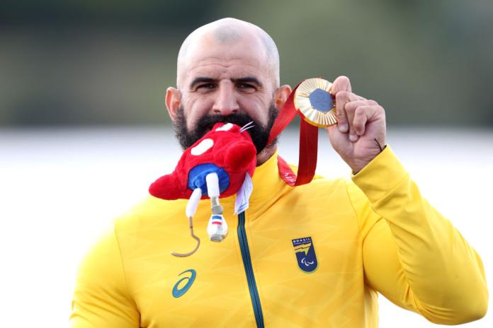 A male Para canoe athlete wearing a yellow uniform is holding a gold medal and a red Paris 2024 mascot plush